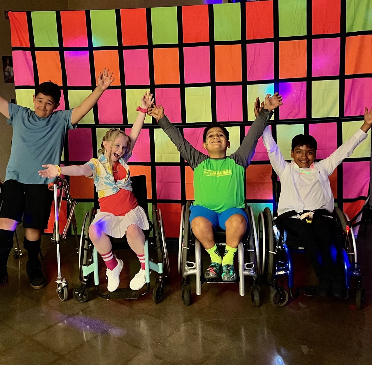 Four kids pose in front of a color background for a picture, with their hands raised.