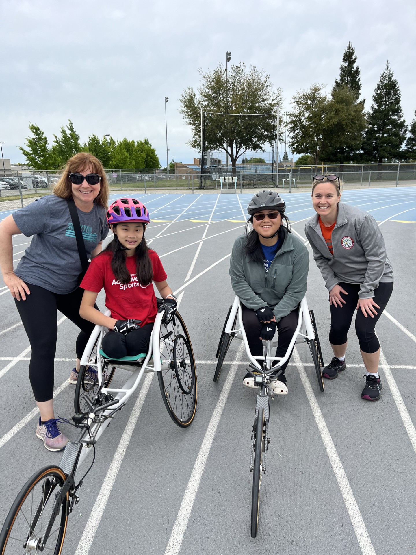 Two athletes sit in their racing chairs on a track, alongside their coaches, posing for a photo.