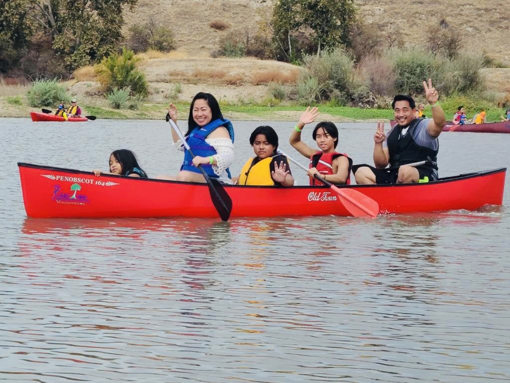 3 kids and two adults paddle a red canoe across water.