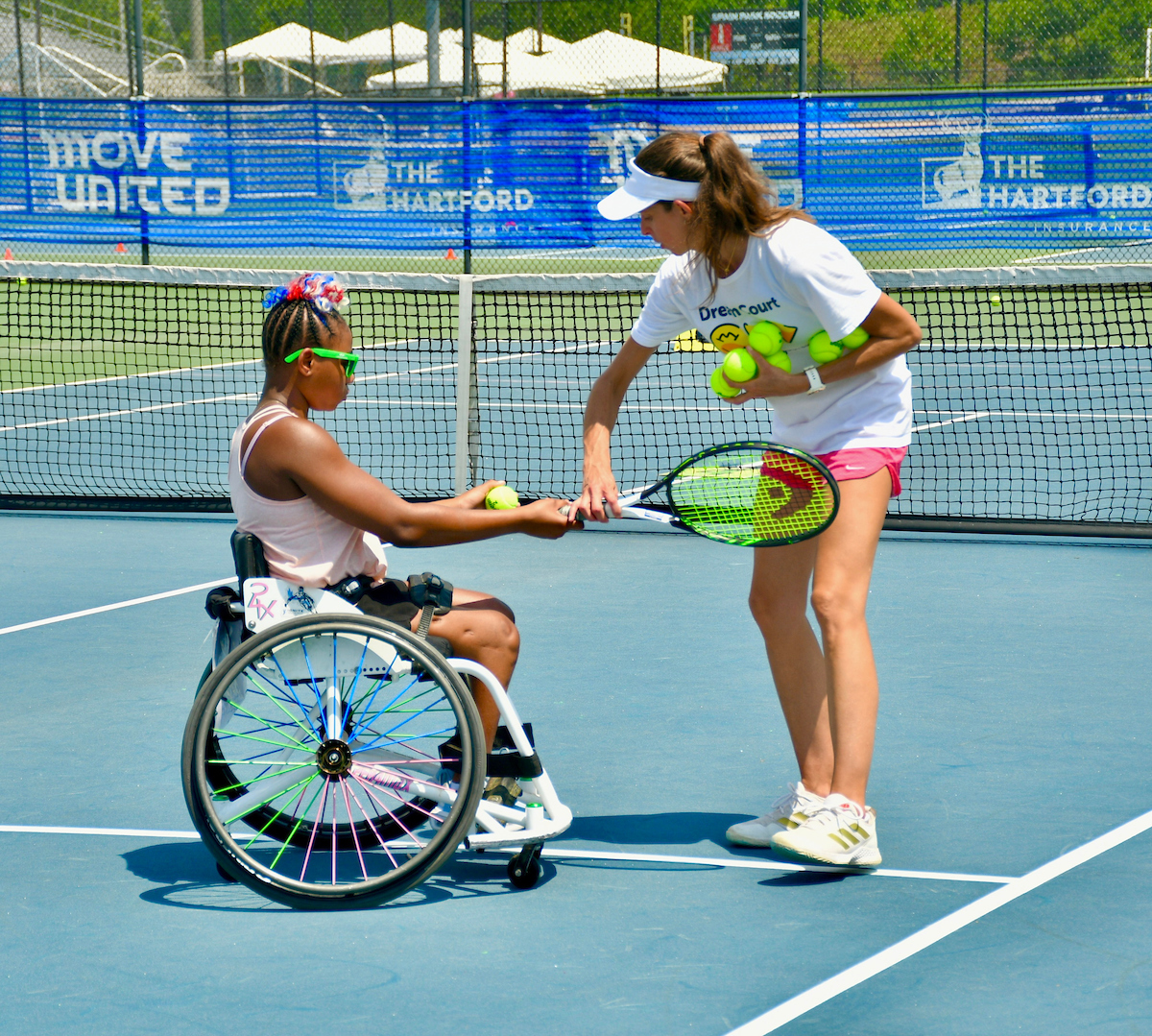 Wheelchair athlete holding racket and balls. Standing volunteer next to athlete adjusting racket in athlete's hand.