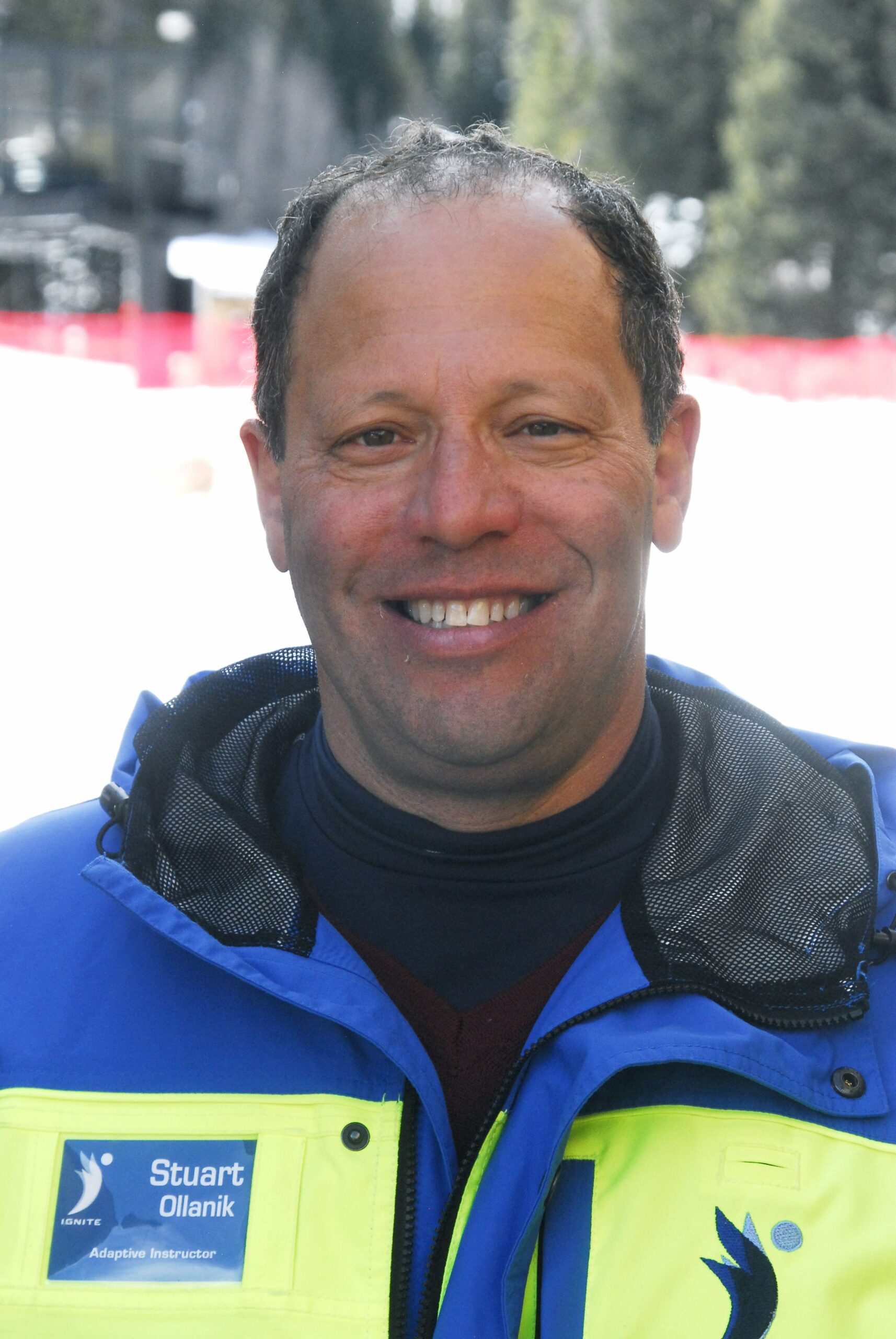 A middle-aged man with short, curly hair smiles at the camera. He is wearing a blue and yellow adaptive instructor jacket with a name badge that reads "Stuart Ollanik." The background is blurred but shows a snowy outdoor setting with red fencing and evergreen trees, suggesting a ski resort or adaptive sports environment.