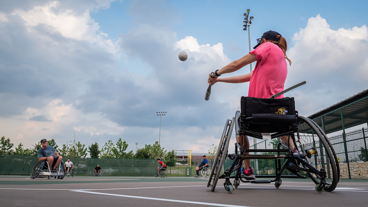 Seated athlete striking softball, softball is headed towards outfield