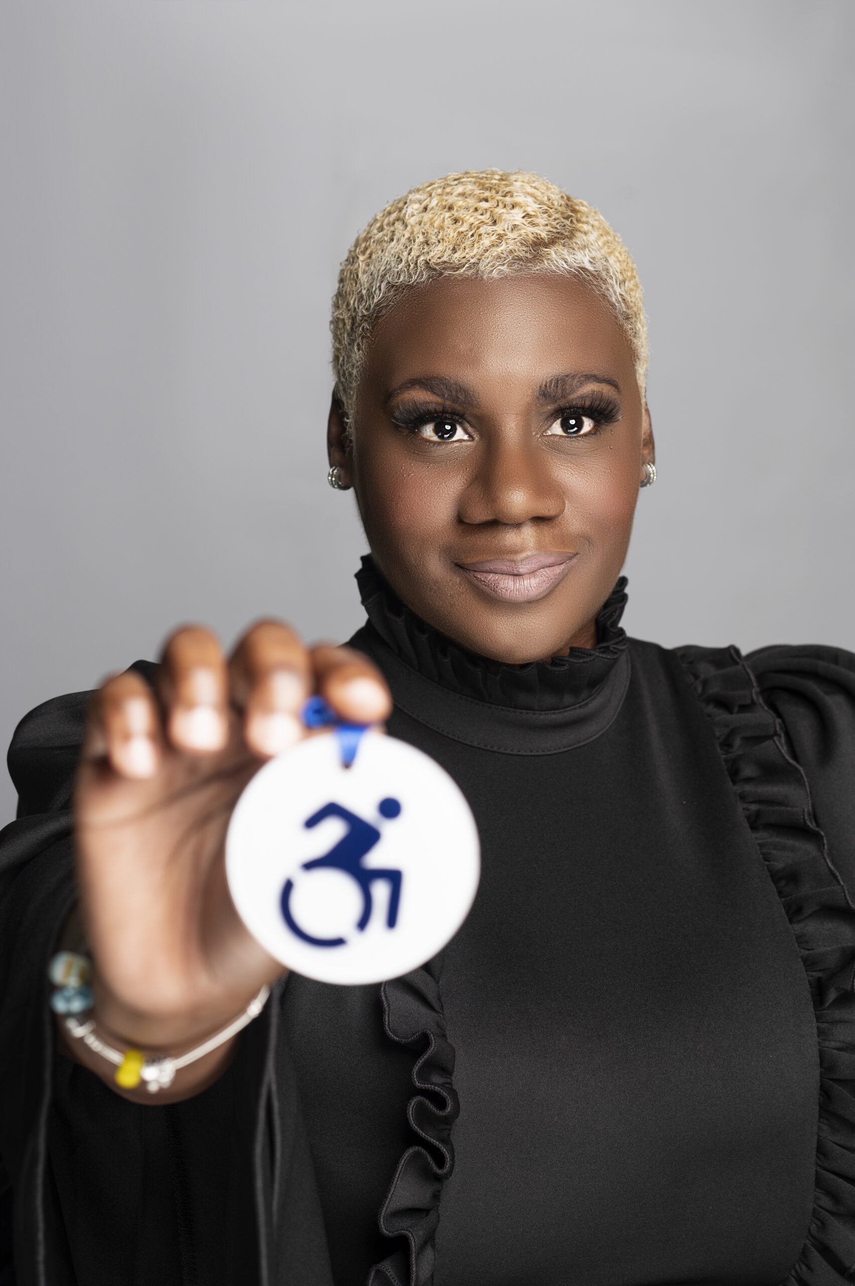 A confident Black woman with short, blonde, curly hair wears a black top with ruffle details and subtle earrings. She holds up a circular accessibility symbol featuring a stylized person in a wheelchair. The focus is on the symbol, slightly blurred in the foreground, while her face is sharply in focus, expressing pride and strength.