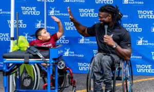 Man in a wheelchair high fives young boy in a wheelchair in front of a backdrop with Move United and The Hartford.