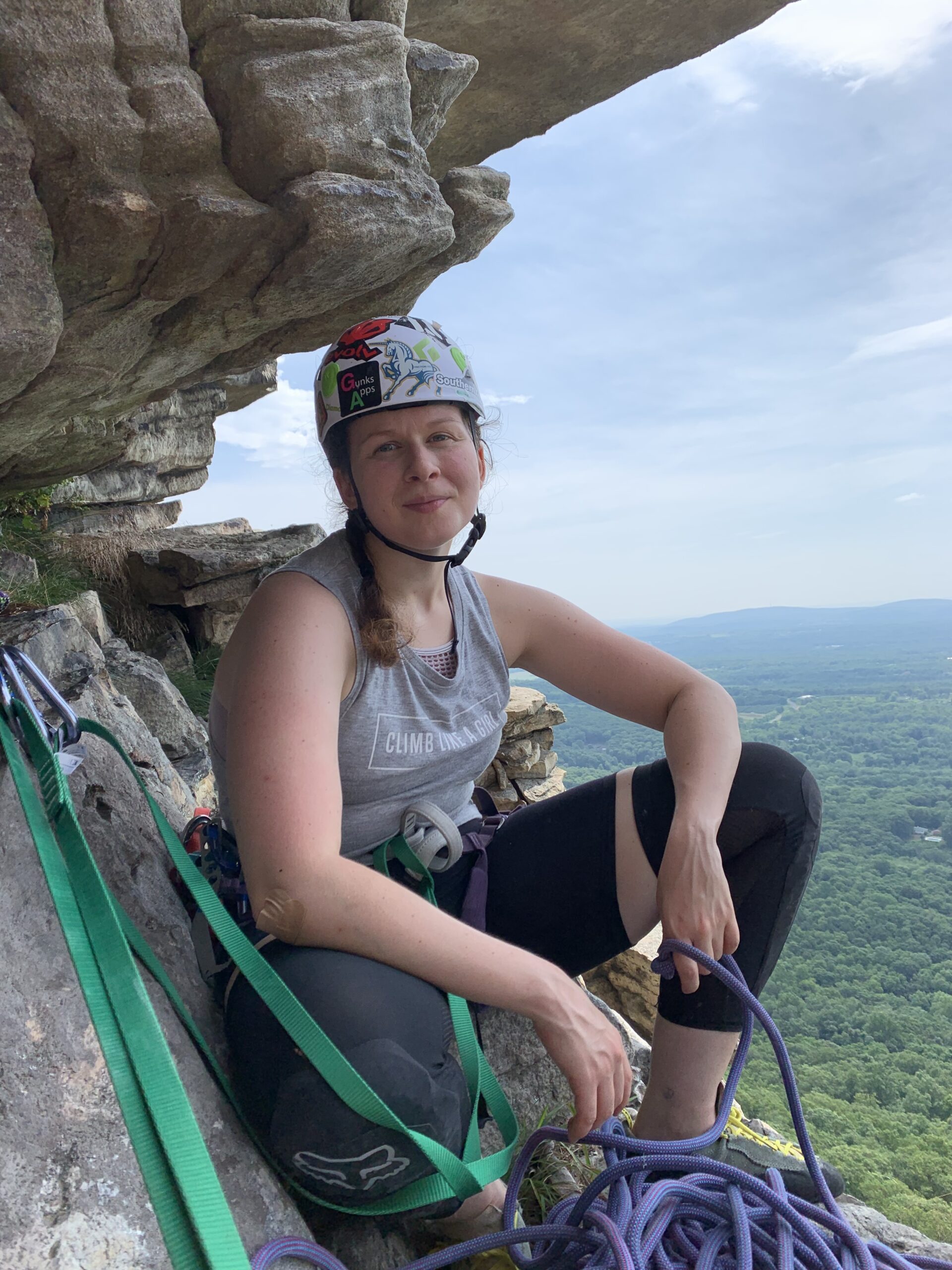A woman wearing a helmet decorated with colorful stickers sits on a rocky ledge partway up a cliff, secured with climbing gear and green safety straps. She wears a sleeveless “Climb Like a Girl” shirt and black leggings, holding a coiled purple rope. Behind her is a vast, scenic view of green forest and distant hills under a blue sky.