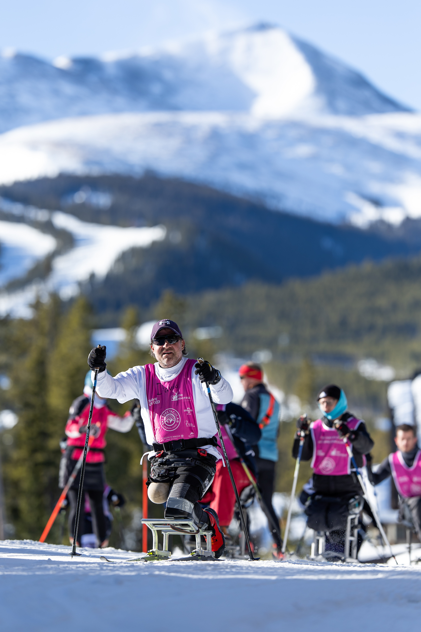 A group of Nordic Sit Skiers going up a hill with the mountains in the background