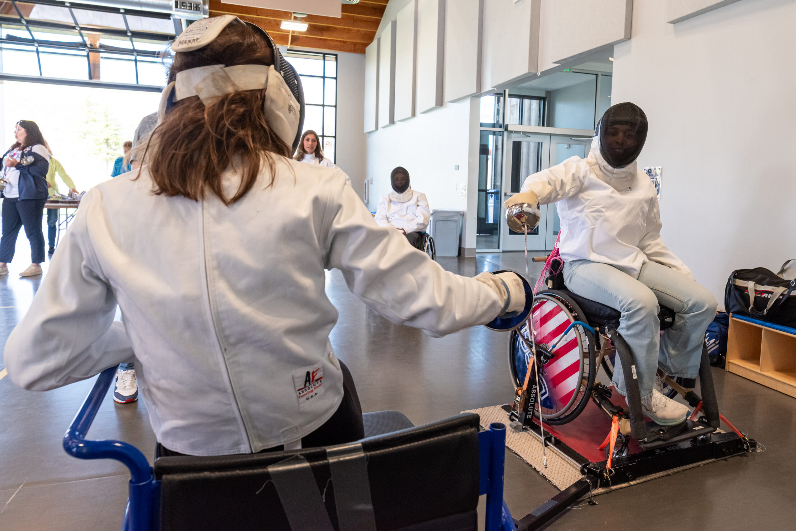 Two seated athletes particpating in a wheelshair fencing practice. They are actively engaged in a match against each other