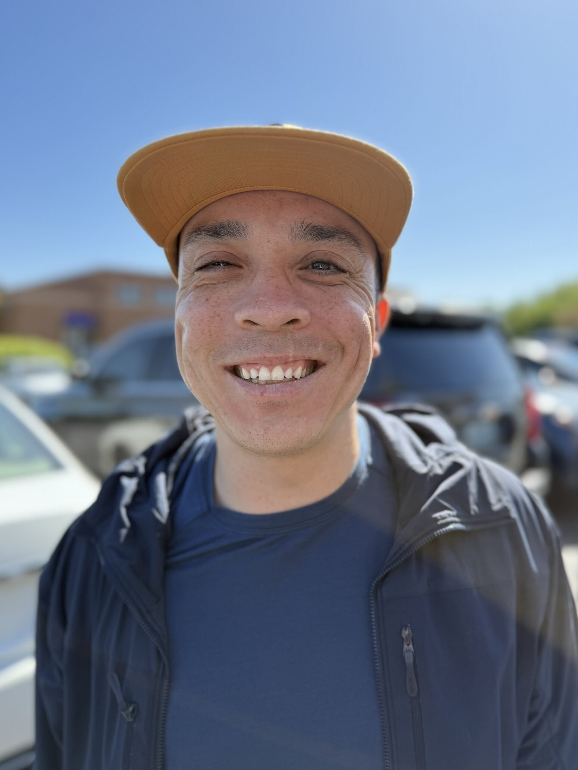 U.S. Navy veteran Jared Lenahan smiling outdoors on a sunny day, wearing a yellow cap, navy shirt, and dark jacket with cars and buildings in the background.