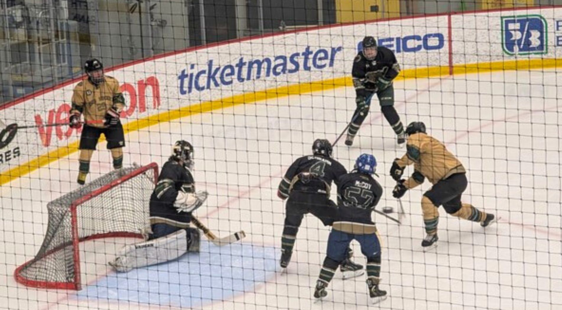 A blind hockey game in action on the ice. A player in a gold jersey prepares to take a shot toward the net while defenders in dark jerseys and the goalie position themselves to block. Another gold team player stands near the goal, and several players cluster around the puck in front of the net, showcasing teamwork and intensity in the adapted sport.