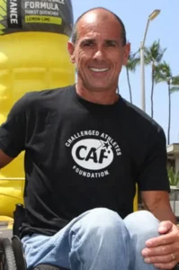 Carlos Moleda in a black t shirt, blue jeans, smiling at the camera while sitting in his wheelchair.