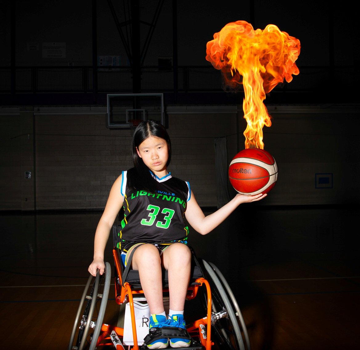 Move United Member Athlete Ella Willis, a young wheelchair basketball player wearing a black “Lightning” jersey with the number 33, poses on a gym court while holding a basketball in one hand. The ball has been edited to appear as if it is on fire, creating a dramatic, energetic effect.