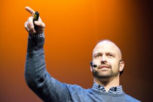 Chirs Kaag speaking at an even in front of a faintly orange background. He has a microphone wrapped around his head and is pointing into the crowd.