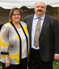 Debbie and Ralph Amernto. Debbie wear a yellow cardigan with thinly drawn black circles and Ralph wears a black suit with a blue undershirt and tie.