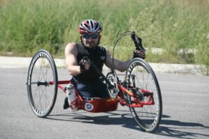 Troy Davis on his adaptive bike wearing a black vest, black sunglasses, and a red, white and blue racing helmet.