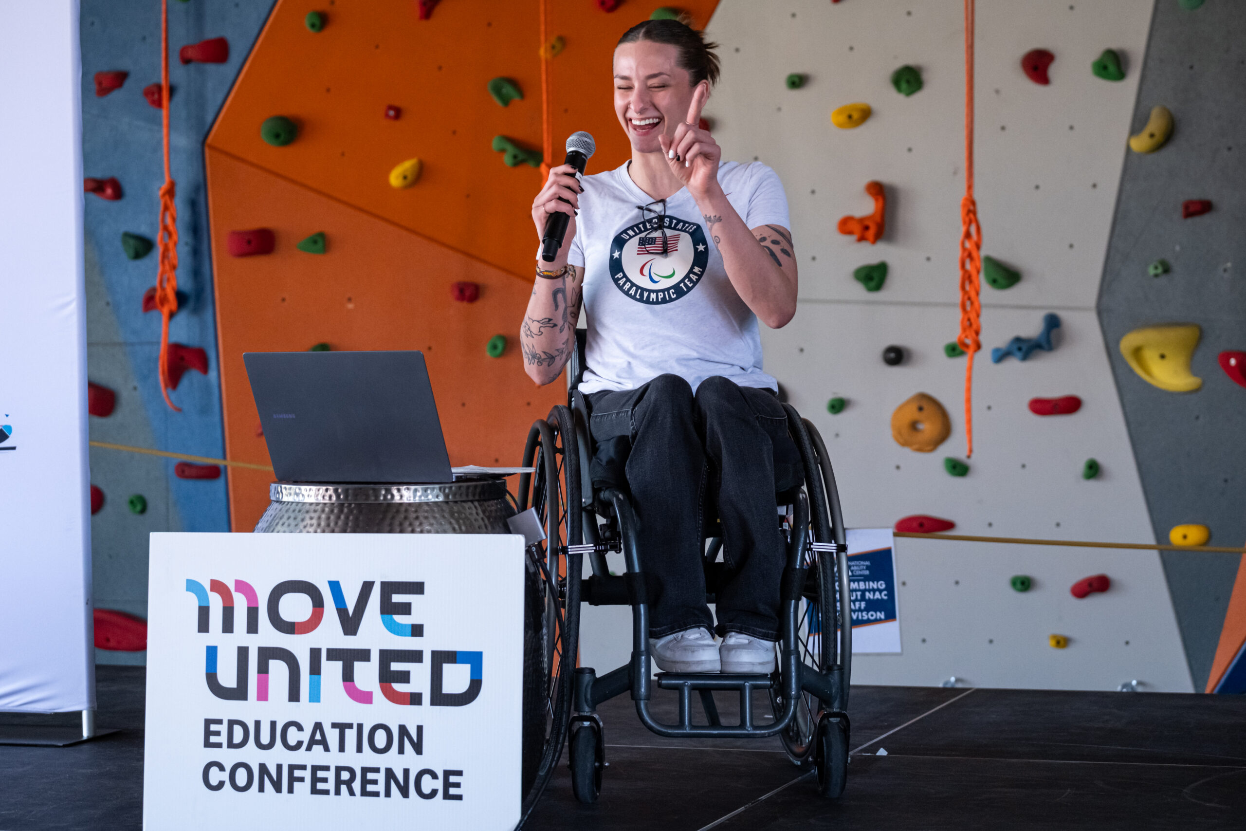 Keynote speaker who is a wheelchair user on stage with Move United Education Conference sign in front of her and rock wall behind her. Speaker is smiling with microphone to mouth and hand in the air making an enthusiastic gesture
