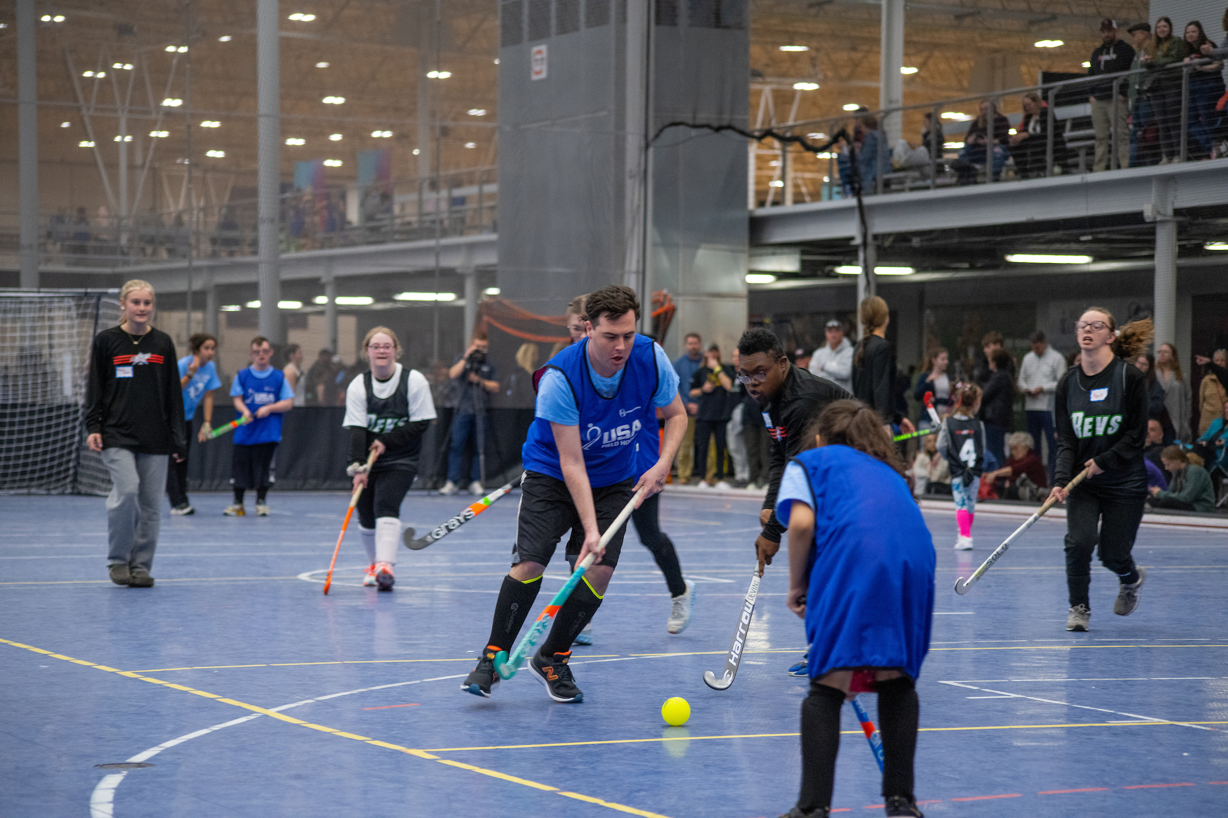 Athlete goes to hit ball with field hockey stick while another athlete plays defense.