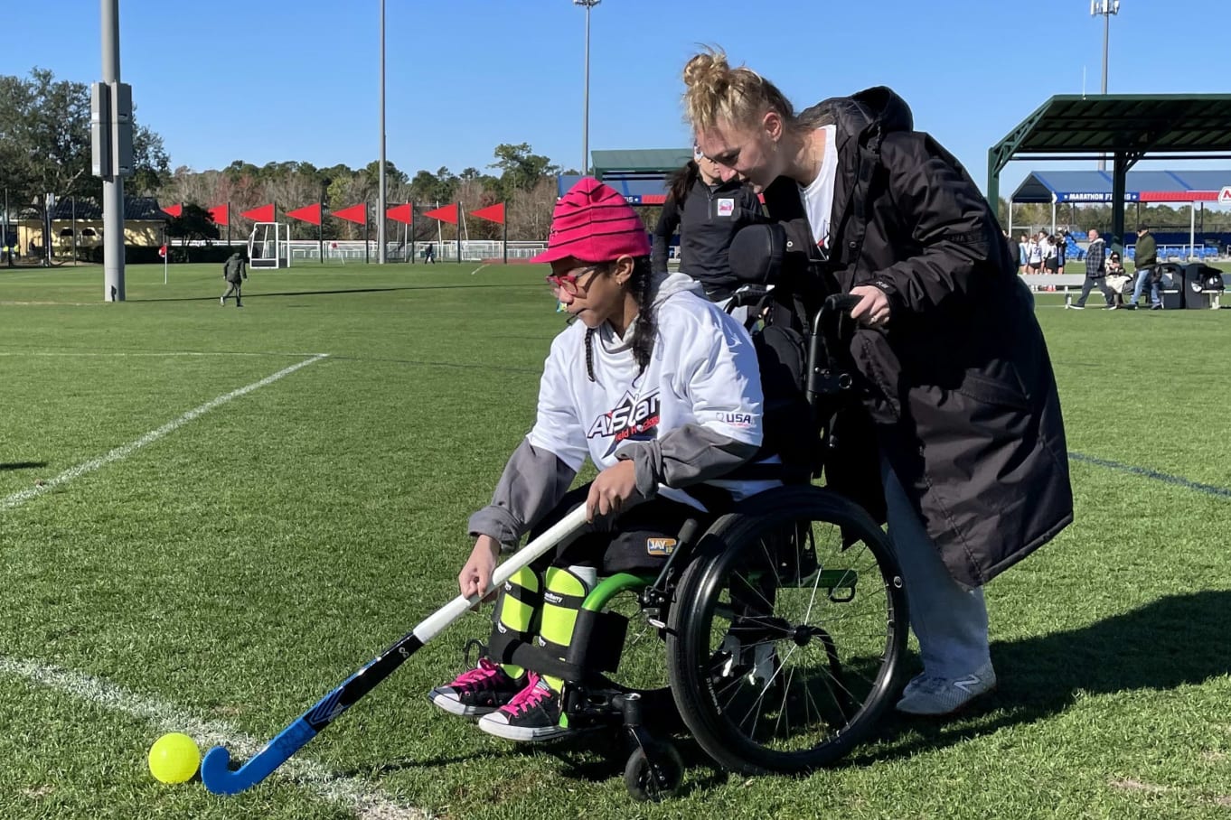 A young athlete in a wheelchair uses a field hockey stick to hit a bright yellow GO ball during an adaptive field hockey session.