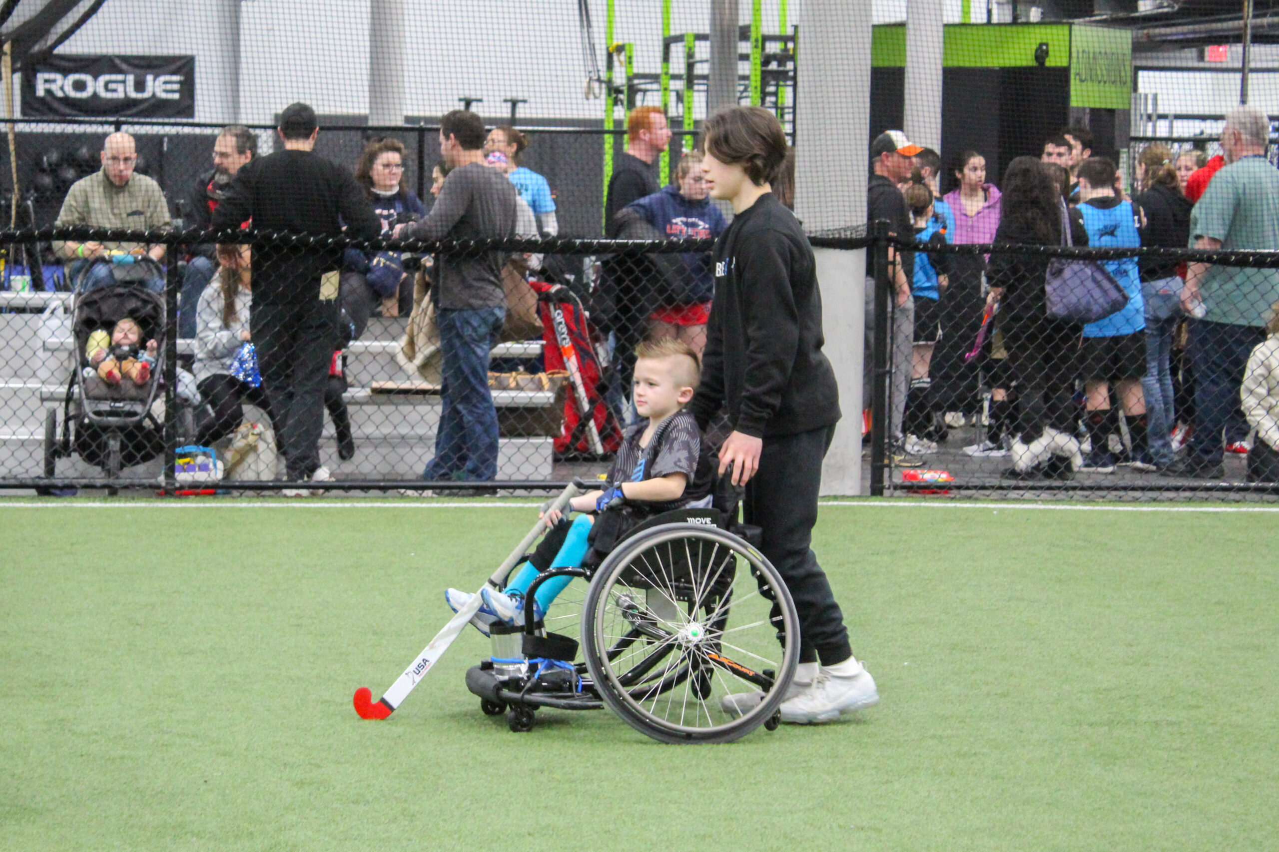 Individual pushing a child athlete in a wheelchair who is playing field hockey