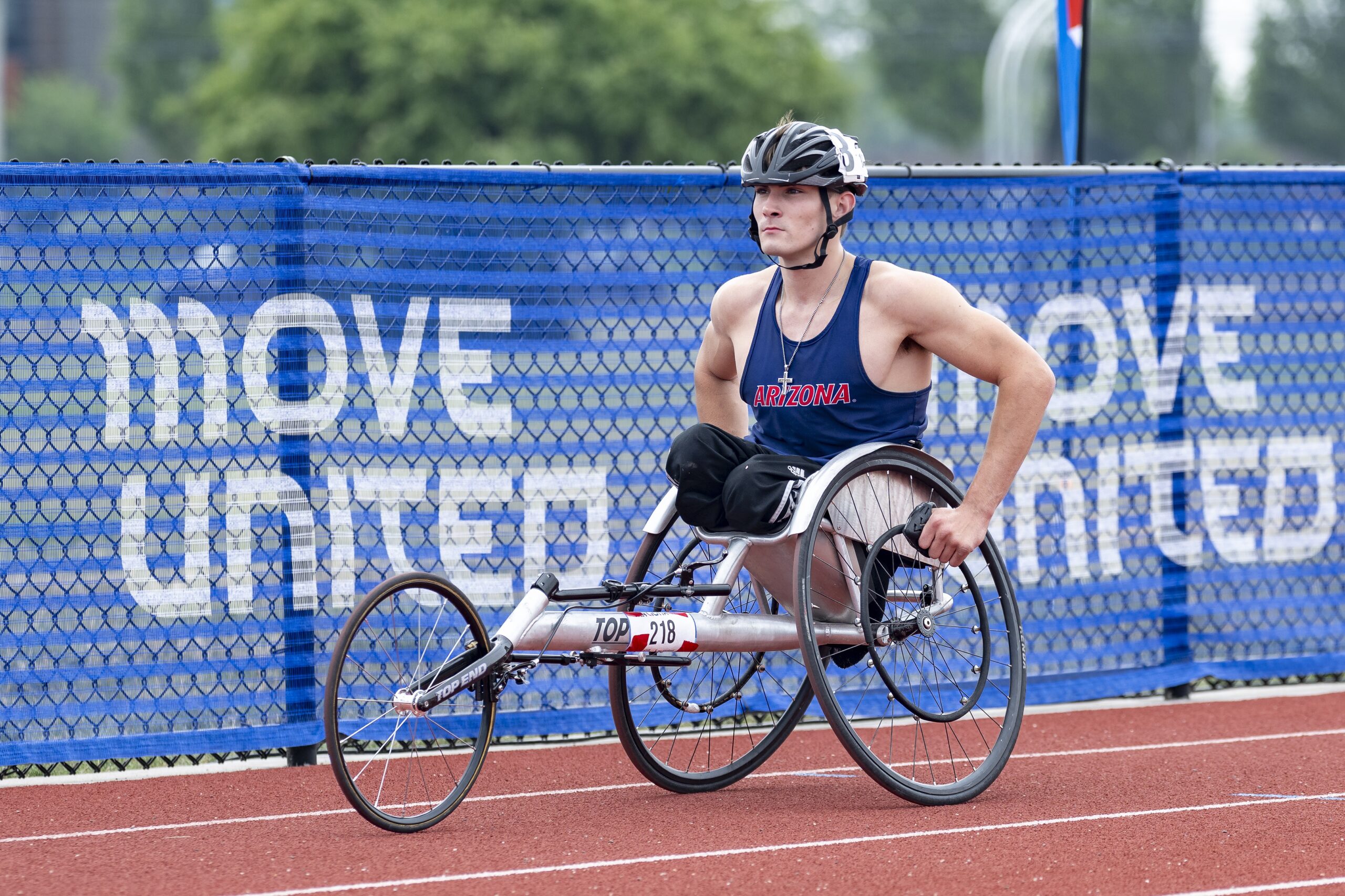 Double below knee amputee athlete in Arizona racing jersey in racing chair on track with with Move United fencing in background