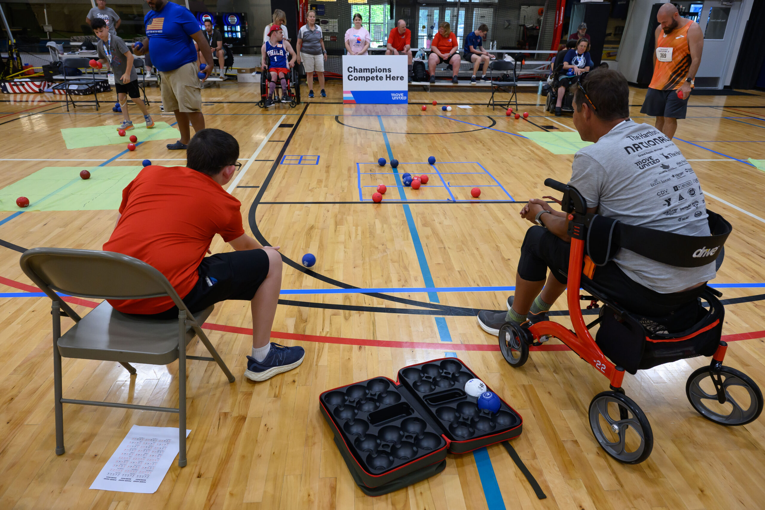 Participant sitting in chair releasing boccia ball towards a taped grid of 9 squares. Another participant seated on walker beside him. In the background, several athletes and participants are spectating this exercise