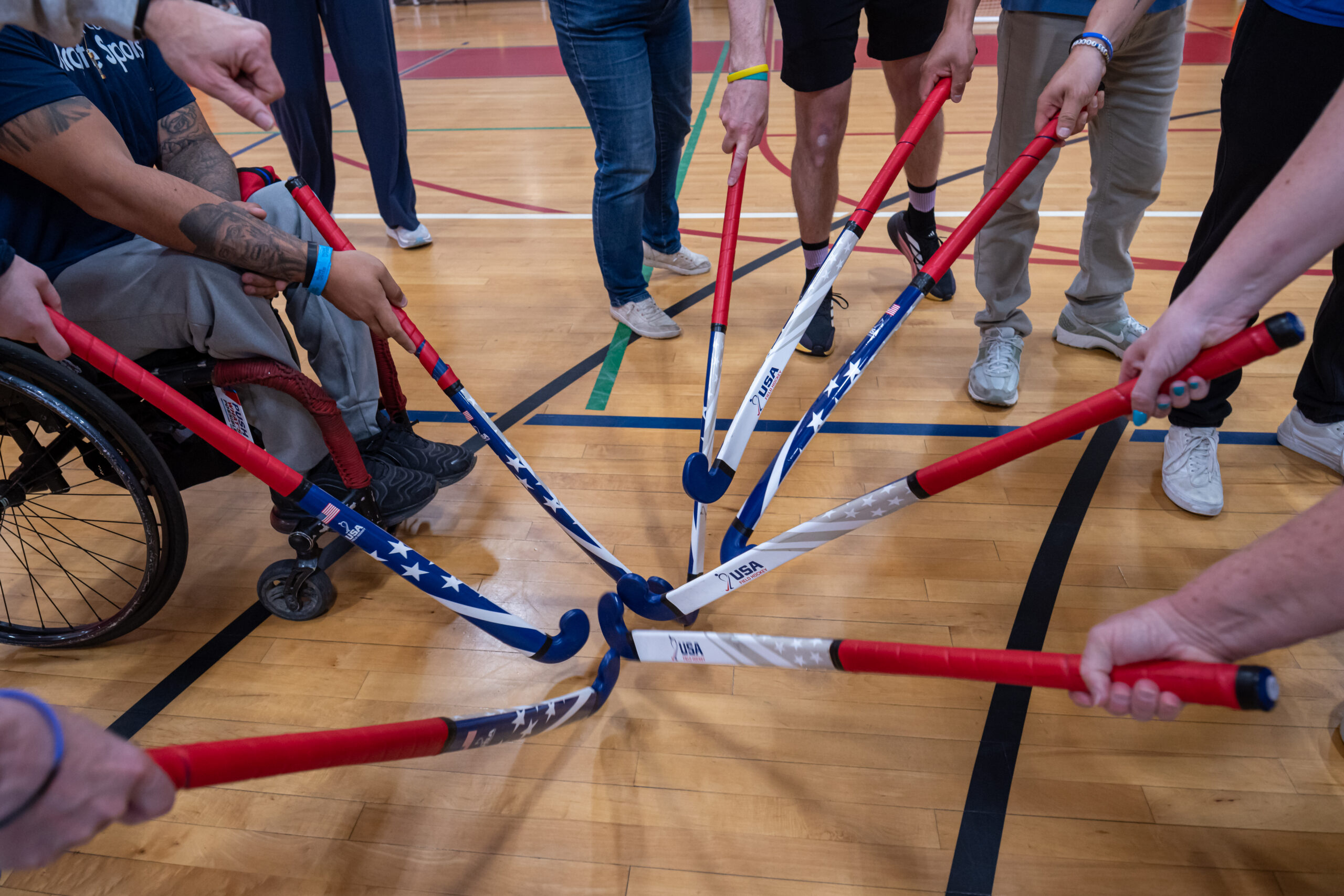 Group of adaptive field hockey session attendees gathered in circle pointing field hockey sticks toward center of circle