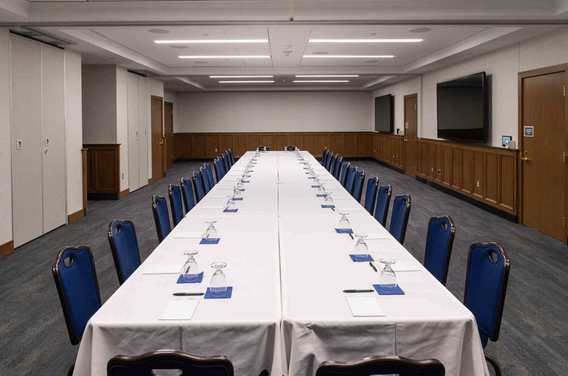 Large breakout room with one long horizontal table with a white tablecloth and 15 chairs on each side.