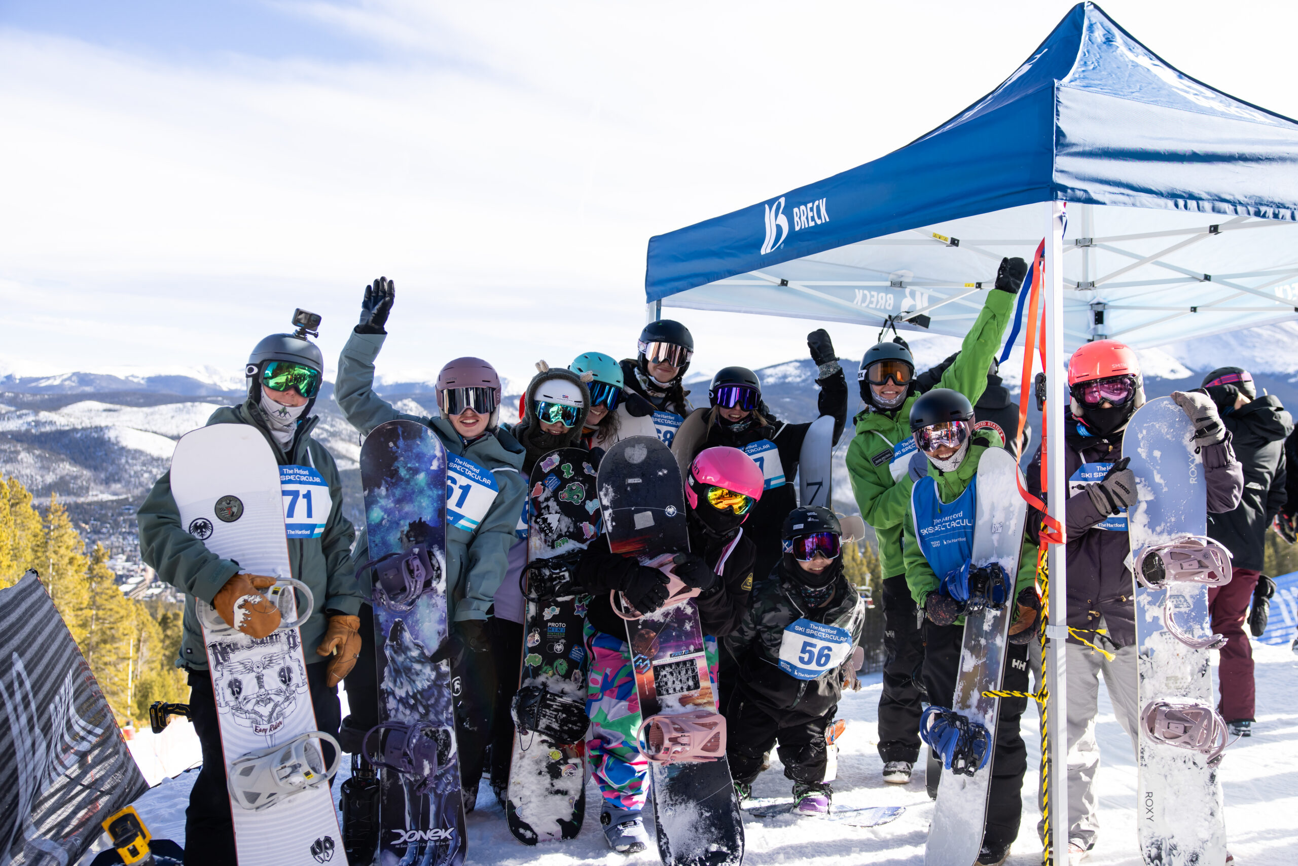 A group of standing snowboarders pose at the top of the mountain next to a Breck tent. All athletes are showing happiness and excitement