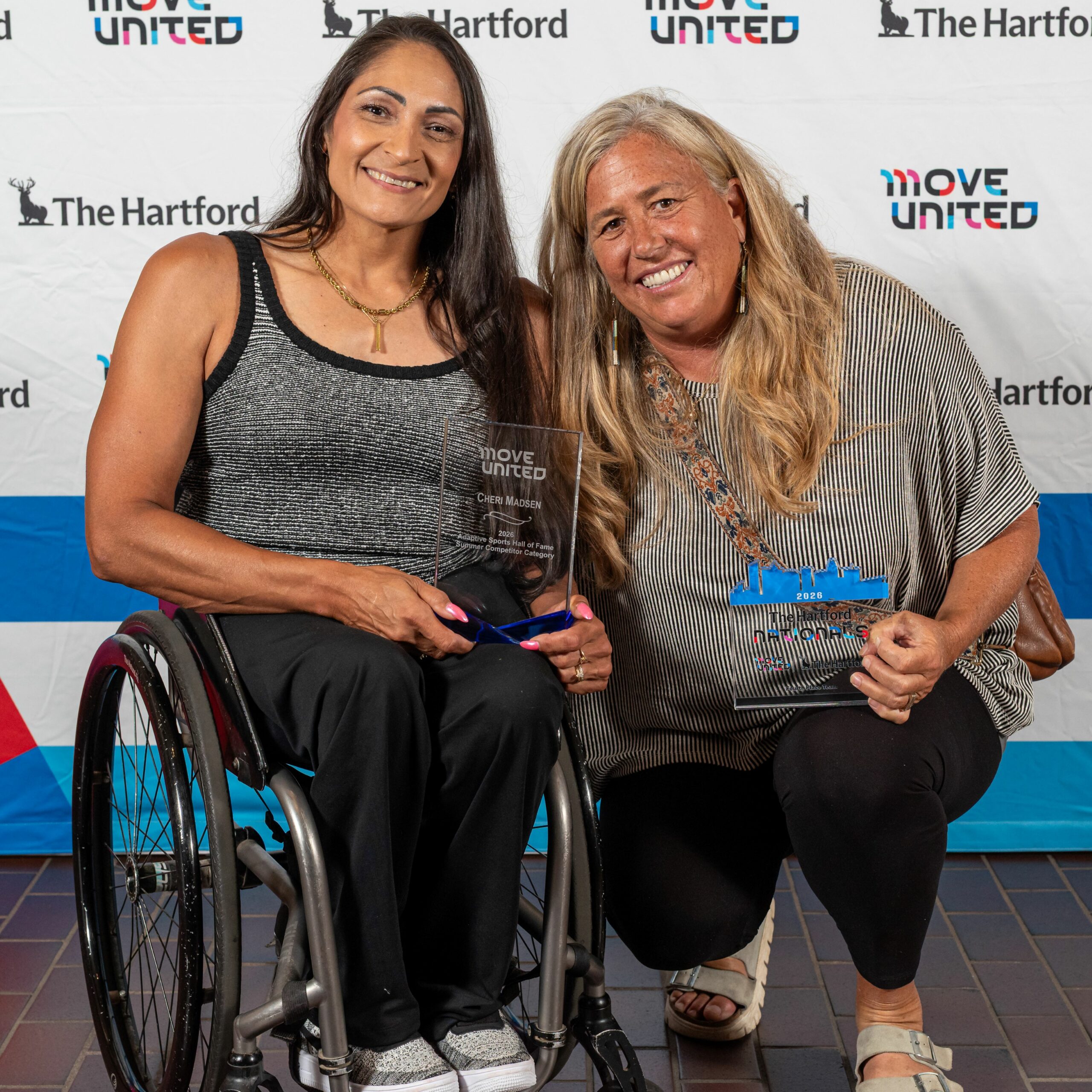 Cheri Madsen and Wendy Gumbert pose with awards in front of a Move United backdrop