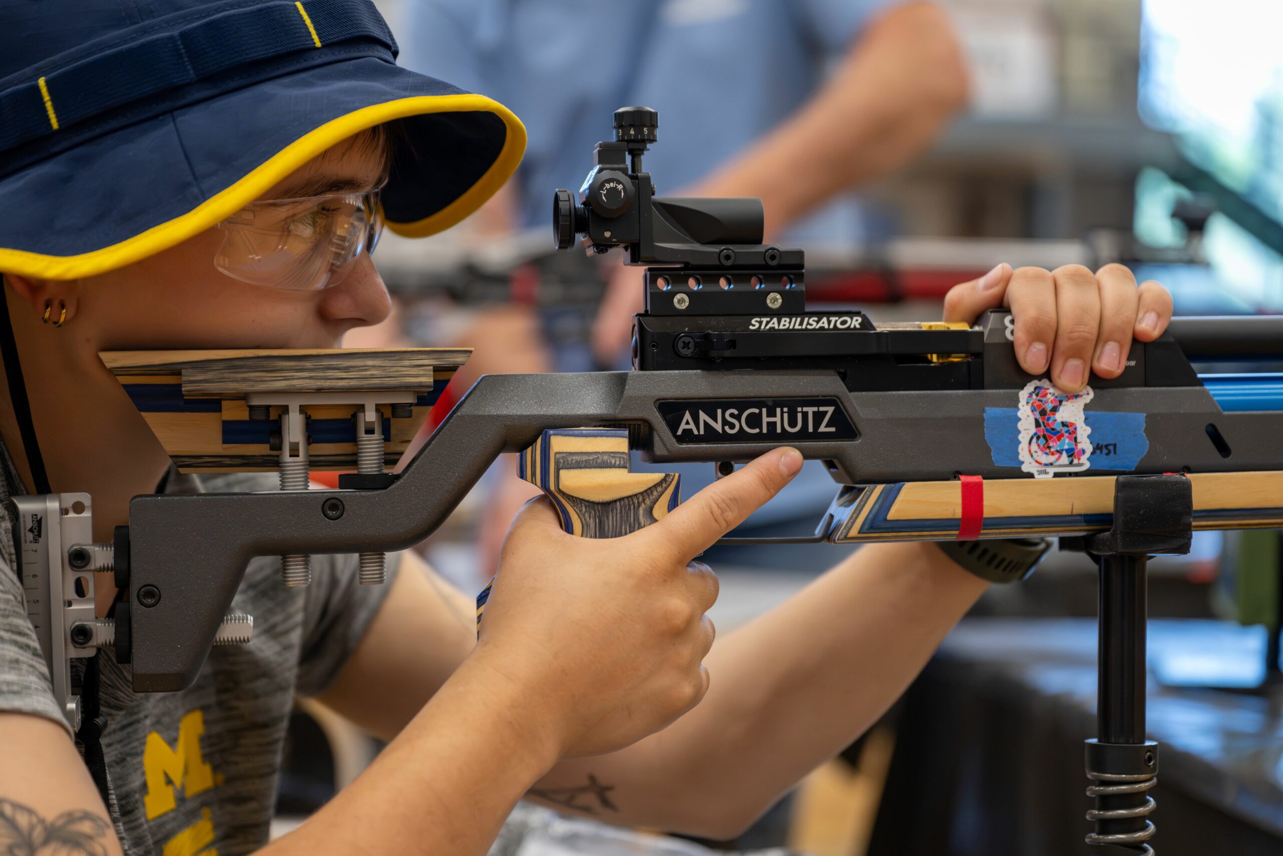Athlete wearing University of Michigan shirt with air rifle set on stand. Athlete's finger is getting ready to pull the trigger.